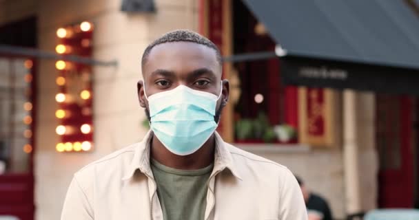 Портрет афроамериканского молодого красивого мужчины в медицинской маске, смотрящего в камеру и стоящего на улице возле кафе. Крупный план красивого мужчины на открытом воздухе. Пандемия коронавируса. — стоковое видео