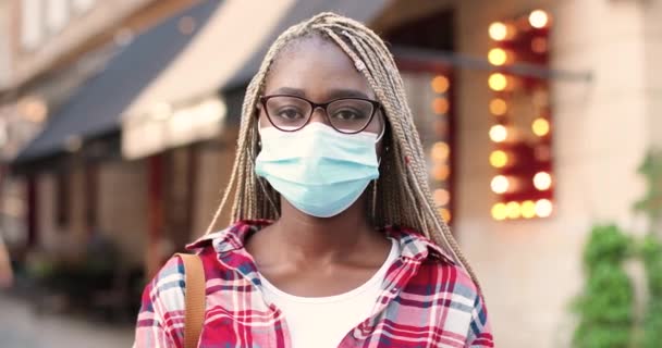 Portretul unei tinere afro-americane în mască medicală și ochelari care se uită la cameră și stau pe stradă lângă cafenea. Aproape de frumoasa femeie în aer liber. Conceptul de coronavirus pandemic . — Videoclip de stoc