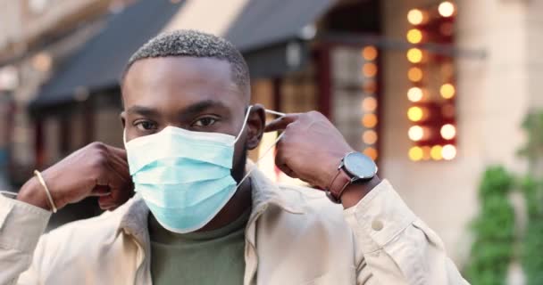 카메라 앞에서 웃고 길거리에 서서 의료용 마스크를 벗은 아프리카계 미국인 젊은이의 모습. 야외에 잘 생긴 수컷을 클로즈업 합니다. 유행성 코로나 바이러스 개념. — 비디오