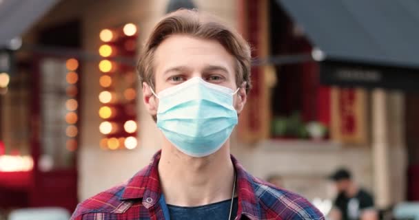 Портрет кавказького молодого чоловіка в медичній масці, який дивиться на камеру і стоїть на вулиці біля кафе. Вигляд чоловіка на вулиці. Пандемічний коронавірус.. — стокове відео