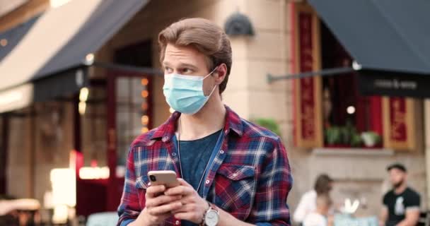 Молодий кавказький стиліст в медичній масці стоїть на вулиці і пише повідомлення на смартфоні. Поручні чоловіки стукають і прокручують мобільний телефон у кафе в місті. Зовні. — стокове відео