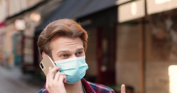 Jonge blanke stijlvolle man met een medisch masker die op straat loopt en praat op een mobieltje. Knappe man die op de mobiele telefoon spreekt en rondloopt. Buiten. Telefoongesprek. — Stockvideo