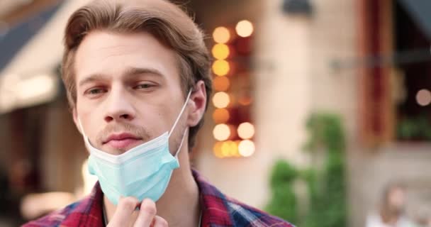 Ritratto di giovane bell'uomo caucasico sorridente alla telecamera, in piedi in strada e togliersi la maschera medica. Primo piano di bel maschio all'aperto. Concetto di coronavirus pandemico. — Video Stock