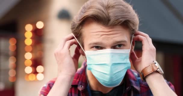 Κοντινό πλάνο του καυκάσιου νεαρού όμορφου άντρα που φοράει ιατρική μάσκα και κοιτάζει την κάμερα στο δρόμο κοντά στο καφέ. Πορτρέτο του άνδρα σε εξωτερικούς χώρους φορώντας αναπνευστική προστασία. Πανδημική έννοια. — Αρχείο Βίντεο