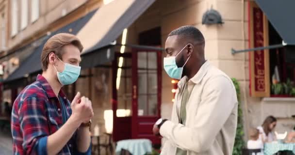 Mixed-races young males friends in medical masks meeting at street, greeting with elbows and talking. African American and Caucasian men at street in pandemic. Multi ethnic guys at social distance. — Stock Video