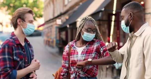 Молоді друзі в медичних масках зустрічаються на вулиці, вітаються ліктями і розмовляють. Африканські кавказькі чоловіки і жінка на вулиці під час пандемії. Багатонаціональні люди на відстані. — стокове відео