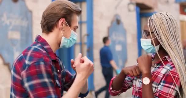 Jovens e alegres amigos em máscaras reunidos na rua, cumprimentando com cotovelos e conversando. Homem e mulher afro-americanos e caucasianos na cidade. Casal multi étnico à distância social. — Vídeo de Stock