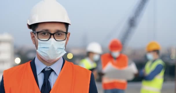Portrét kavkazského pohledného mladého stavitele v kazajce, brýlích a lékařské masce, stojícího venku na stavbě a hledícího do kamery. Close up of businessman investor at building in helmet. — Stock video
