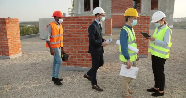 Donna caucasica in casco e maschera medica in piedi a costruire lato e lasciando entrare i lavoratori maschi di razze miste al mattino, controllando la temperatura. Costruttori e ingegneri multietnici nell'edilizia. — Video Stock