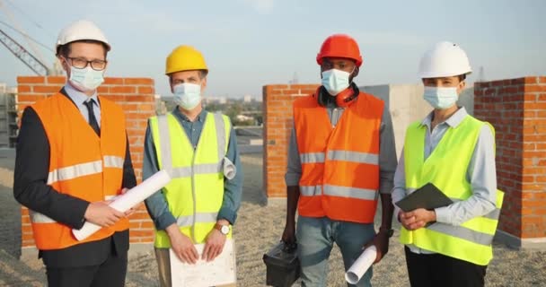 Portret van gemengde rassen mannen en vrouwen constructeurs team in helmen en medische maskers staan op het bouwen van top met het bouwen van plannen ontwerpen. Coronavirus. Ingenieurs en architecten in de bouw. — Stockvideo