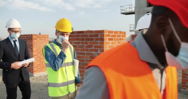 Mujer caucásica en máscara médica que mide y comprueba la temperatura en los hombres trabajadores mixtos de la construcción. Multi hombres étnicos constructores e ingenieros que vienen en la construcción. Coronavirus. — Vídeos de Stock