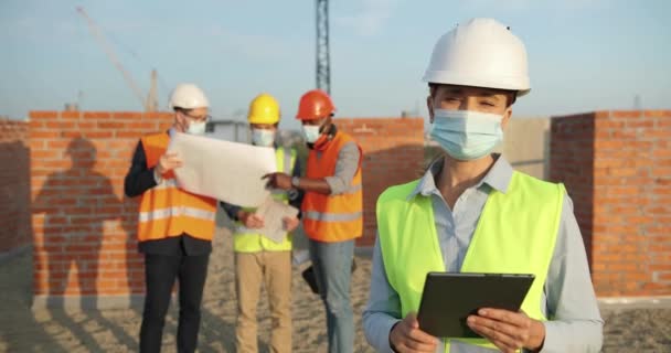 Porträt einer kaukasischen Bauarbeiterin in Kaskade und medizinischer Maske, die draußen auf der Baustelle steht. Bauarbeiterin oder Ingenieurin beim Bauen in Helm mit Tablet-Gerät in der Hand. Coronavirus-Pandemie — Stockvideo