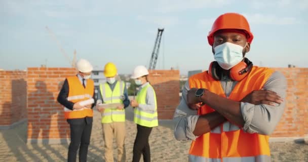 Retrato do jovem construtor afro-americano bonito em casco e máscara médica em pé ao ar livre na construção e olhando para a câmera. Construtor masculino no topo da construção em capacete. Coronavírus. — Vídeo de Stock