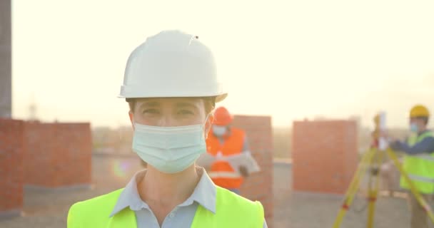 Retrato caucásico joven constructor femenino en casco y máscara médica de pie al aire libre en la construcción. Primer plano de la mujer constructora en el edificio en casco. Coronavirus. Constructores trabajando en segundo plano. — Vídeo de stock