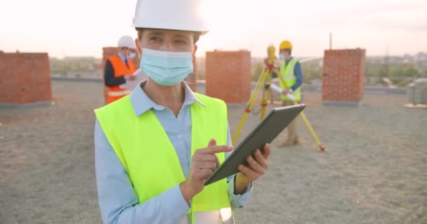 Mujer blanca joven y guapa ingeniera en casco y máscara médica de pie en la construcción de lado, utilizando el dispositivo de la tableta y tocando en la pantalla. Al aire libre. Constructor femenino con ordenador gadget. — Vídeo de stock