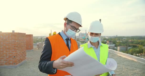 Kaukasiska par manliga och kvinnliga konstruktörer i hjälmar och medicinska masker talar och diskuterar planutkast på hög byggnadssida. Man och kvinna byggare arbetar på att bygga. Pandemi. — Stockvideo