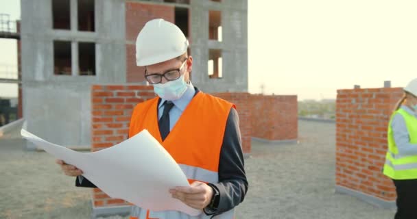 Caucásico joven empresario inversor en hemet y máscara médica de pie en el edificio alto y mirando proyecto de plan de construcción en las manos. Ingeniero o arquitecto masculino en la construcción de lado en pandemia. — Vídeo de stock