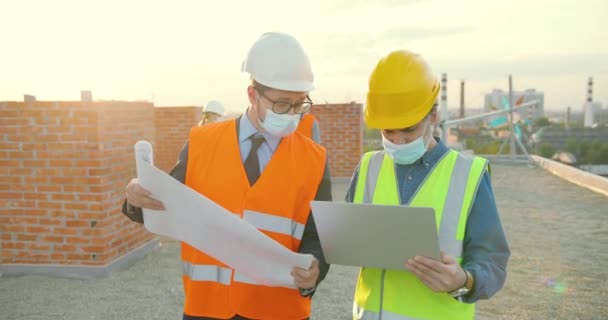 Hombres caucásicos constructores colegas en cascos y máscaras médicas hablando y discutiendo proyecto de plan en el lado de la construcción. Los hombres constructores que trabajan en la construcción con el ordenador portátil. Socios. — Vídeo de stock