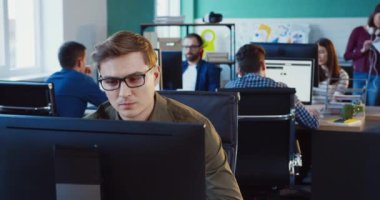 İş yerinde otururken bilgisayar ekranına bakan gözlüklü yakışıklı bir adamın portresi. Arkaplan ofis çalışanları.