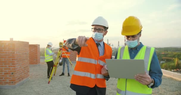 Bělošský podnikatel investor dává příkazy k nákupu nebo inženýr s notebookem a ukazuje prstem. Konstrukční strana. Konstruktoři v lékařských maskách a helmách diskutující o stavbě. — Stock video