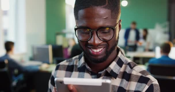 Primer plano retrato de un joven afroamericano con camisa a cuadros y gafas. Trabajador de oficina sonriendo sosteniendo la tableta en sus manos. — Vídeos de Stock