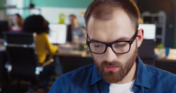 Προσωπογραφία νεαρού Καυκάσου με γένια σε γυαλιά. Όμορφος υπάλληλος γραφείου με τζιν πουκάμισο κοιτάζει την κάμερα.. — Αρχείο Βίντεο