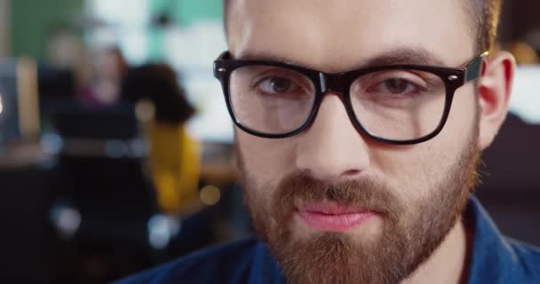 Primer plano retrato de un joven caucásico con barba y gafas. Hombre guapo con ojos marrones mirando a la cámara y sonriendo. — Vídeos de Stock