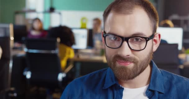 Nahaufnahme Porträt eines jungen kaukasischen Mannes mit Bart und Brille. Schöner Mann im Jeanshemd blickt im Büro in die Kameralinse. — Stockvideo