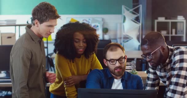 Joven equipo multinacional de cuatro personas discutiendo la solución al problema mientras están de pie en la oficina moderna en la computadora. — Vídeos de Stock
