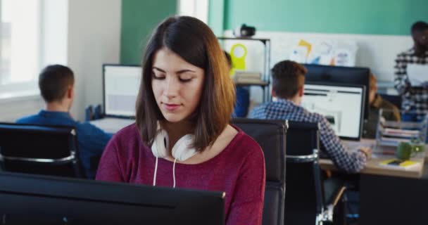 Mladá krásná dívka s bílými sluchátky sedí u počítače a psaní textu s úsměvem. Ženský specialista je rád, že pracuje v kanceláři. — Stock video