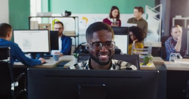 Retrato de un joven afroamericano con gafas trabajando en oficina en computadora. Trabajar como consultor en línea, programador, diseñador o desarrollador en la oficina. — Vídeos de Stock