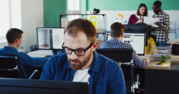 Retrato de hombre joven caucásico en gafas con barba trabajando en la resolución de problemas sentado en la computadora en la oficina. — Vídeo de stock