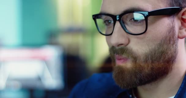 Portret de aproape al unui caucazian chipeș cu barbă în ochelari privind ecranul monitorului și zâmbind . — Videoclip de stoc