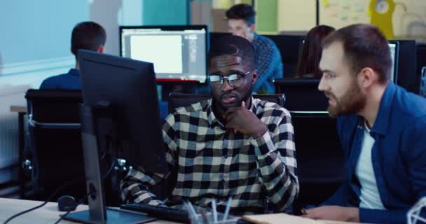 Два сотрудника сидят за компьютером в поисках решения проблемы. Молодой белый мужчина в джинсовой рубашке делает заметки в блокноте. — стоковое видео