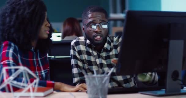 Két afro-amerikai irodai dolgozó ül az asztalnál és nézi a képernyőt. Gyönyörű, göndör hajú nő elmagyarázza az ötletét a kollégájának.. — Stock videók