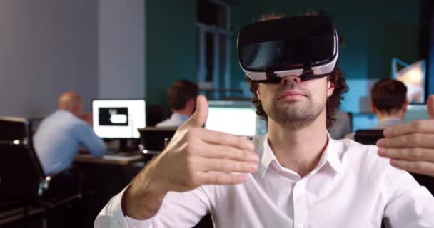 Snygg kontorsarbetare i vit skjorta sitter i virtuell verklighet hjälm vid sitt skrivbord och gester med händerna i luften. — Stockvideo