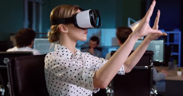 Joven hermosa mujer con el pelo rubio en gestos camisa blanca con las manos sentadas en casco de realidad virtual en la oficina. — Vídeos de Stock