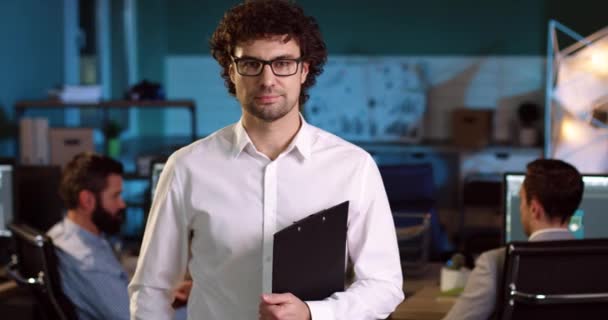 Close up retrato de empresário adulto em camisa branca com óculos de pé no meio do escritório com pasta em suas mãos e sorrindo. — Vídeo de Stock