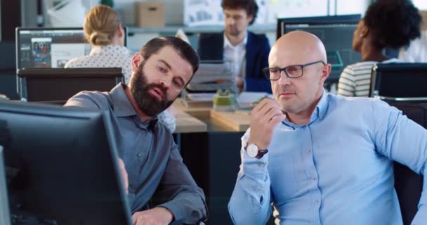 Zwei gut aussehende kaukasische Männer sitzen im Büro am Computer und besprechen die fertige Präsentation des Projekts mit Blick auf den Monitor. — Stockvideo