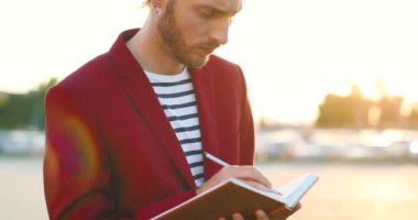 Beyaz, kırmızı ceketli yakışıklı adam planlarını deftere yazıyor. Çekici erkek araba parkında planlama yapıyor. Not bırakmak ve plan yapmak. Arkaplanda arabalar.