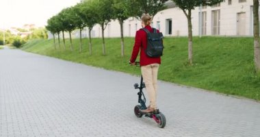 Şehir caddesinde elektrikli scooter kullanan sırt çantalı, beyaz, yakışıklı bir adamın arkası. Modern kasaba. Kırmızı ceketli, şık ve mutlu bir adamın araba sürüşü hakkında arka plan. Erkek sürücü.