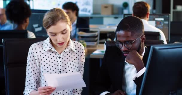 Przystojny młody Afroamerykanin w garniturze i okularach wykonuje zadania siedząc w biurze. Młoda piękna blondynka biznes kobieta sprawdza pracę pracownika i kiwa głową. — Wideo stockowe