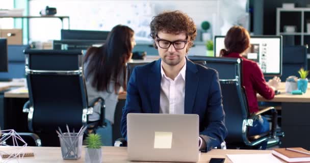 सूट आणि चष्मा मध्ये तरुण कार्यालय कामगार पोर्ट्रेट डेस्कवर बसून ऑफिसमध्ये लॅपटॉपवर काम करीत आहे . — स्टॉक व्हिडिओ