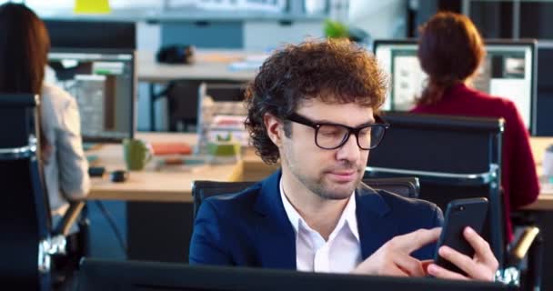 Retrato de funcionário de escritório em óculos e terno sentado no escritório em sua mesa e usando telefone celular. — Vídeo de Stock