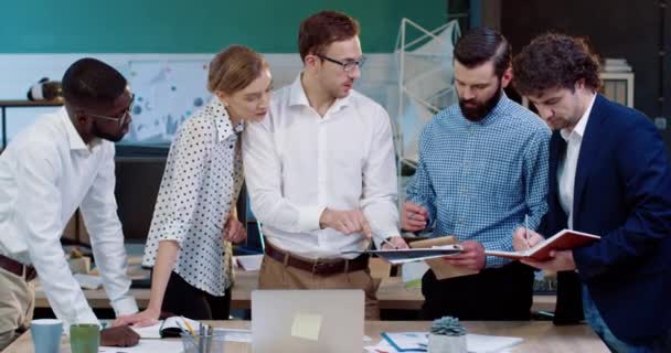 Jonge Kaukasische senior manager in bril geeft taken aan ondergeschikten en toont presentatie op laptop. Kantoorpersoneel noteert. — Stockvideo