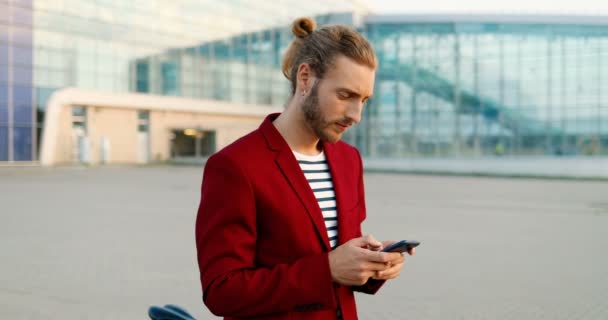 Portrait de beau jeune homme caucasien en veste rouge debout à l'extérieur avec vélo, textos sur smartphone et souriant à la caméra. attrayant heureux mâle tapotement et défilement sur téléphone mobile. — Video