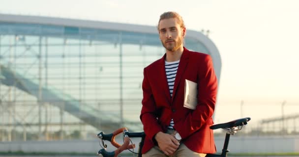 Portret van een knappe, stijlvolle jongeman in een rood jasje, kijkend naar de camera, met een krant onder zijn arm en zittend op de fiets. Mannelijke casual fietser leunend en poseren buiten bij moderne glazen aeroport. — Stockvideo