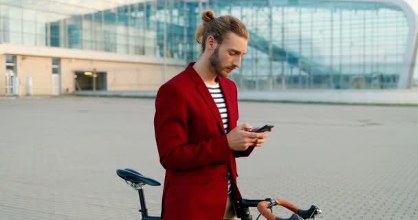Giovane caucasico sorrise bell'uomo in piedi in bicicletta e sms messaggio su smartphone. Elegante ragazzo in giacca rossa appoggiato sulla moto e toccando o scorrendo sul telefono cellulare all'aeroporto. All'aperto. — Video Stock