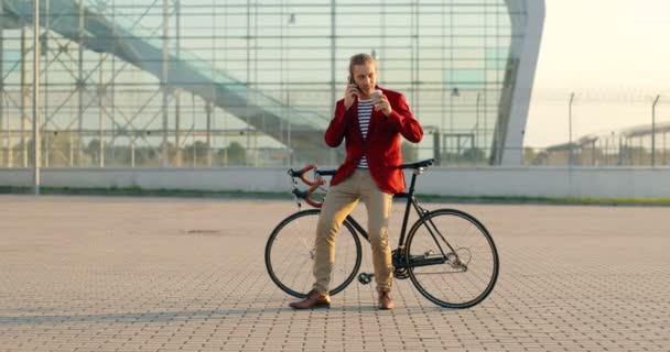 Mannelijke casual knappe fietser in rood jasje zittend op de fiets, nippend aan warme drank en pratend op mobiele telefoon. Bij groot glas modern gebouw. Blanke man die koffie drinkt. en spreken op mobiele telefoon. — Stockvideo