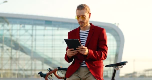 Caucásico joven guapo hombre de pie en bicicleta y el uso de dispositivo de tableta. Chico con estilo en chaqueta roja y gafas de sol apoyadas en la bicicleta y tocando o desplazándose en la computadora gadget en el edificio de vidrio moderno. — Vídeos de Stock